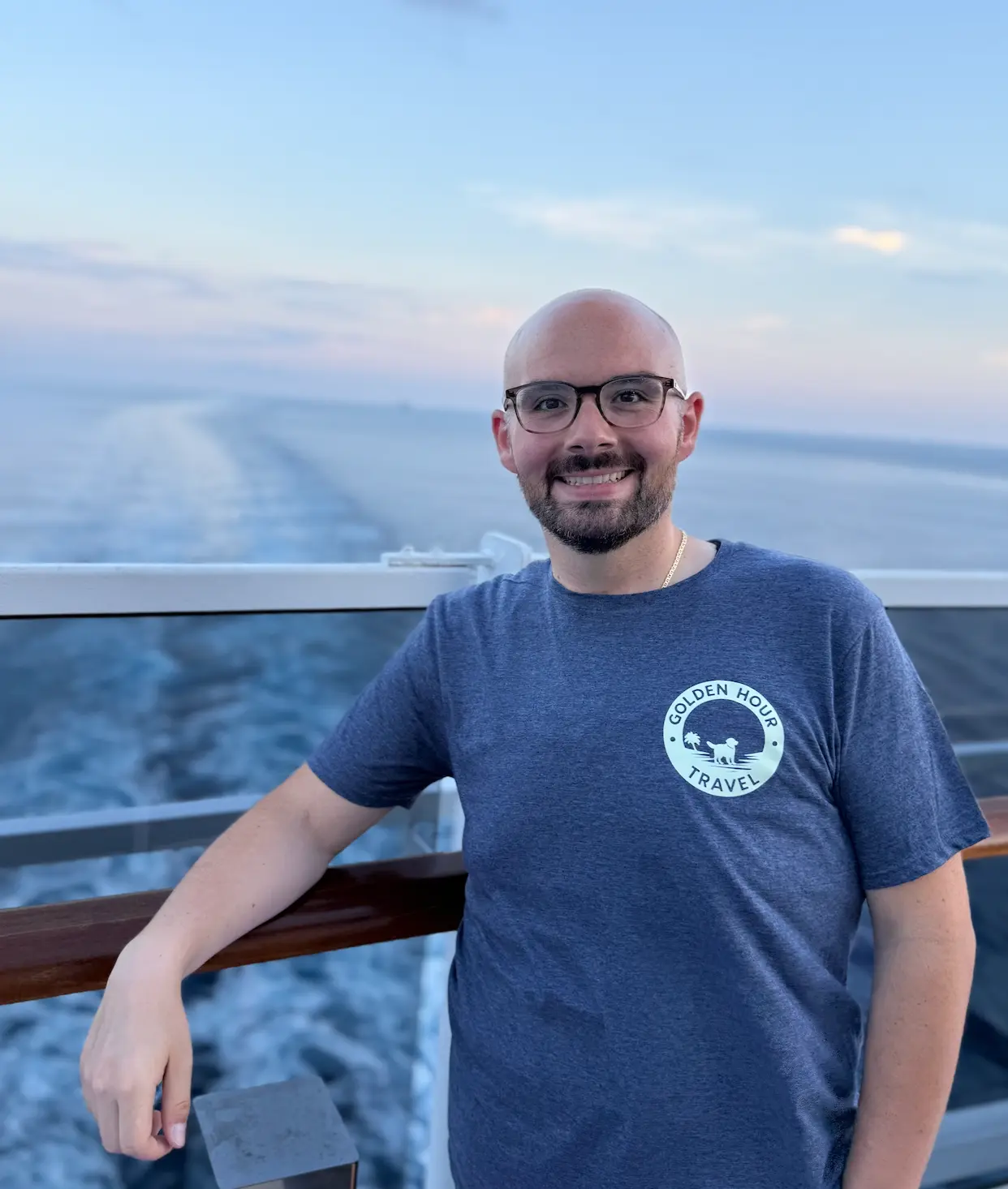 Chris of Golden Hour Travel smiling at a ship's rail in a Golden Hour Travel shirt, with the ocean wake stretching behind him.