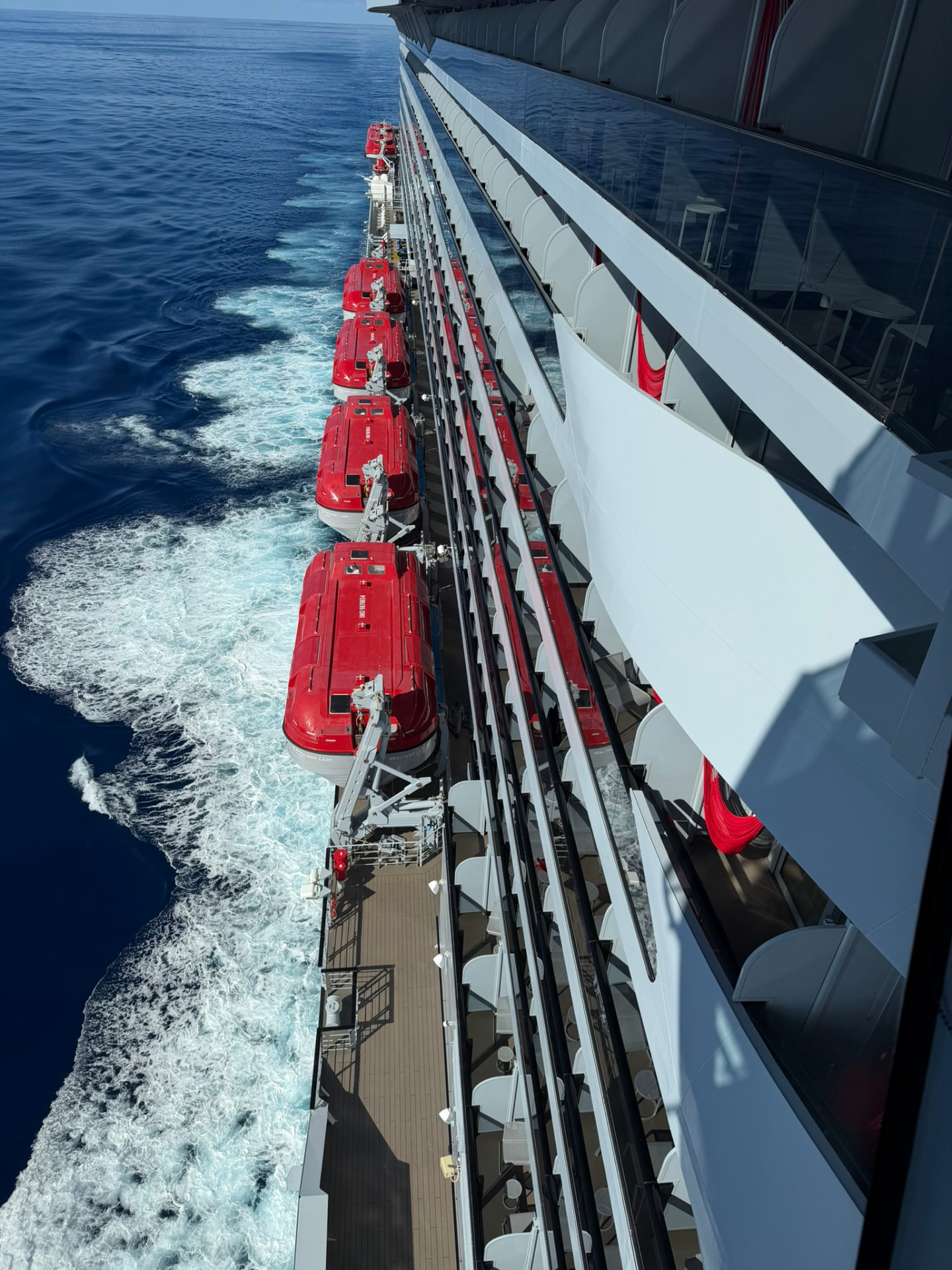 View down the side of a Virgin Voyages ship at sea, with red lifeboats along the deck and the ship's wake in deep blue water.
